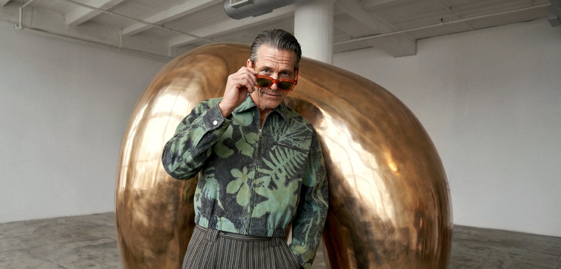 Homme portant une veste fleurie verte et des lunettes de soleil rouges pose devant une grande sculpture de haricot en bronze brillant dans une pièce lumineuse et spacieuse.