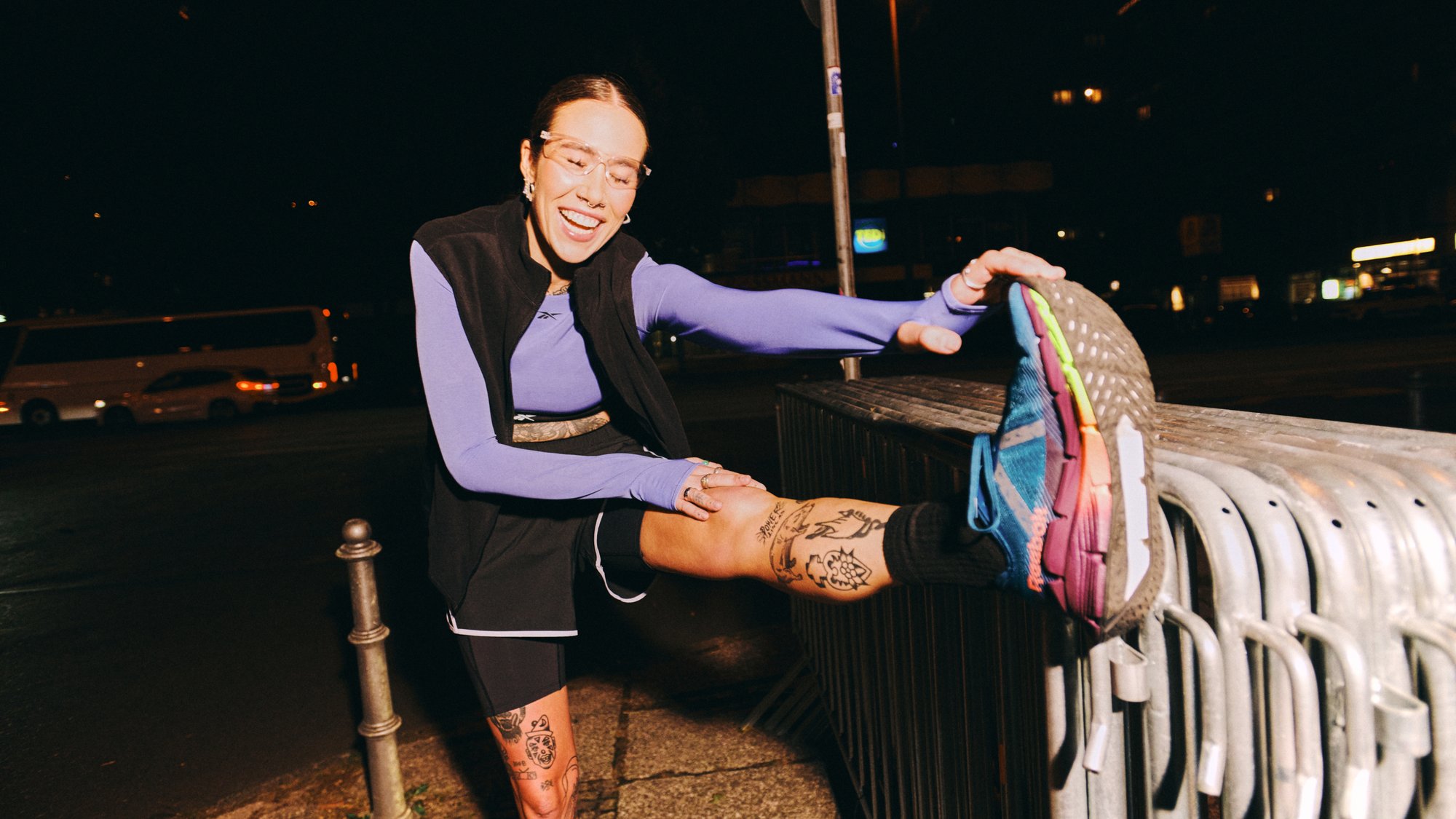 A woman stretches her leg on a railing at night, smiling, wearing athletic gear with visible tattoos on her leg. City lights are in the background.