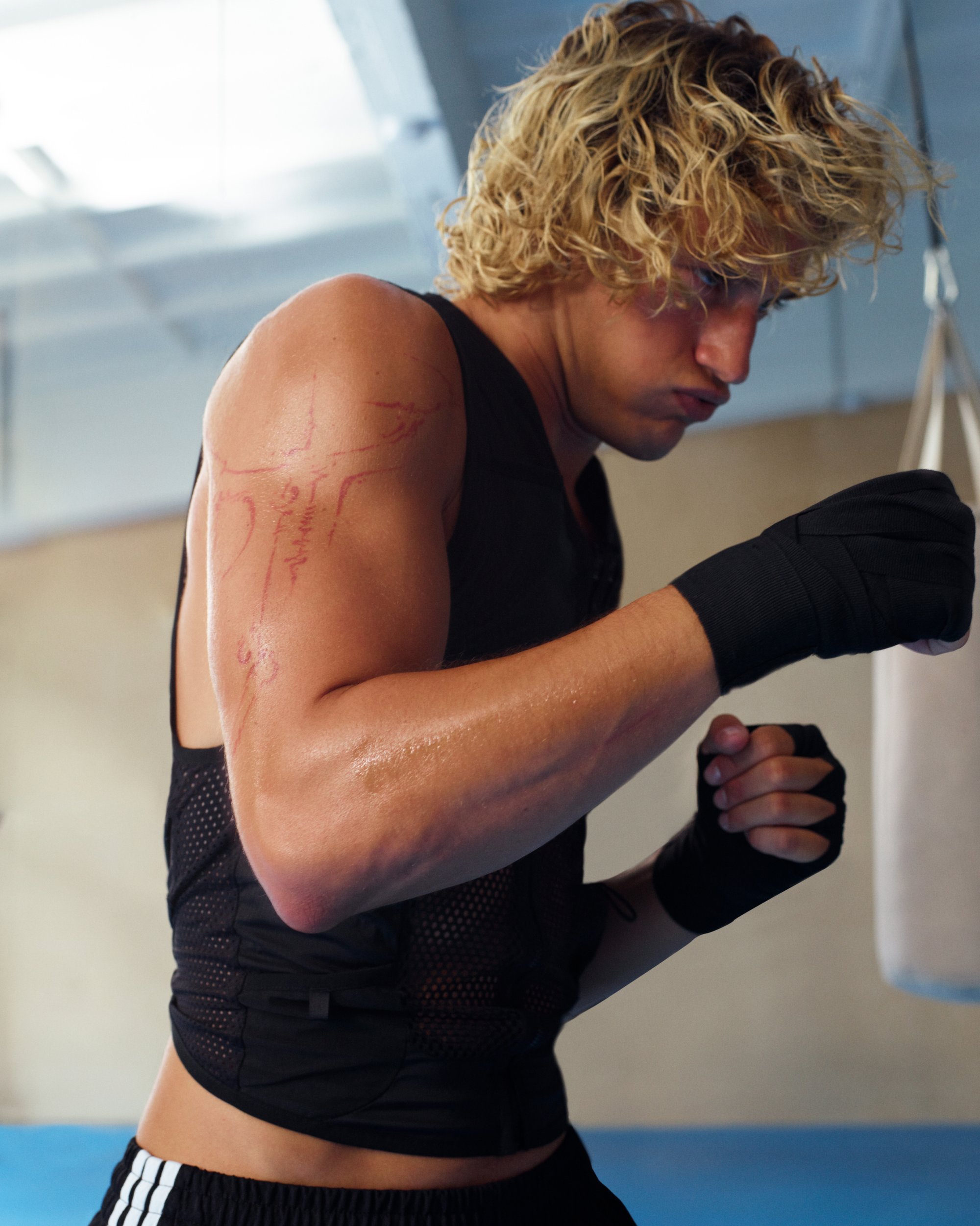 A young man with curly hair, wearing a black athletic top and hand wraps, prepares to punch a hanging punching bag in a gym setting.
