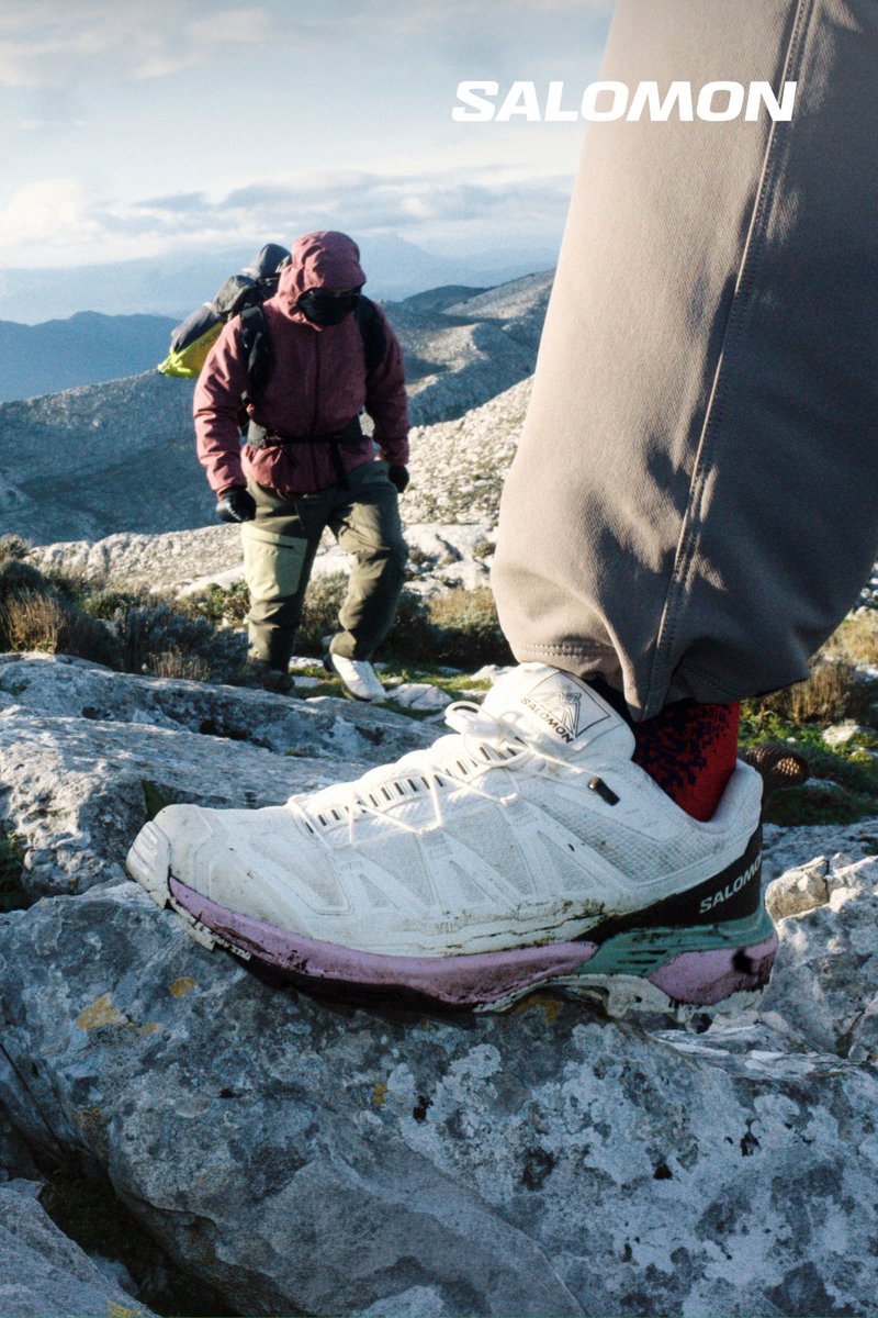 Zapato de senderismo Salomon blanco sobre terreno rocoso con un senderista equipado con ropa de exterior escalando una montaña en el fondo.