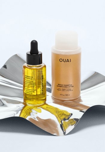 Närande hårbottenolja i en klar flaska med en svart pipett, bredvid en guldschampoflaska med vit kork och svart text, på en reflekterande yta.