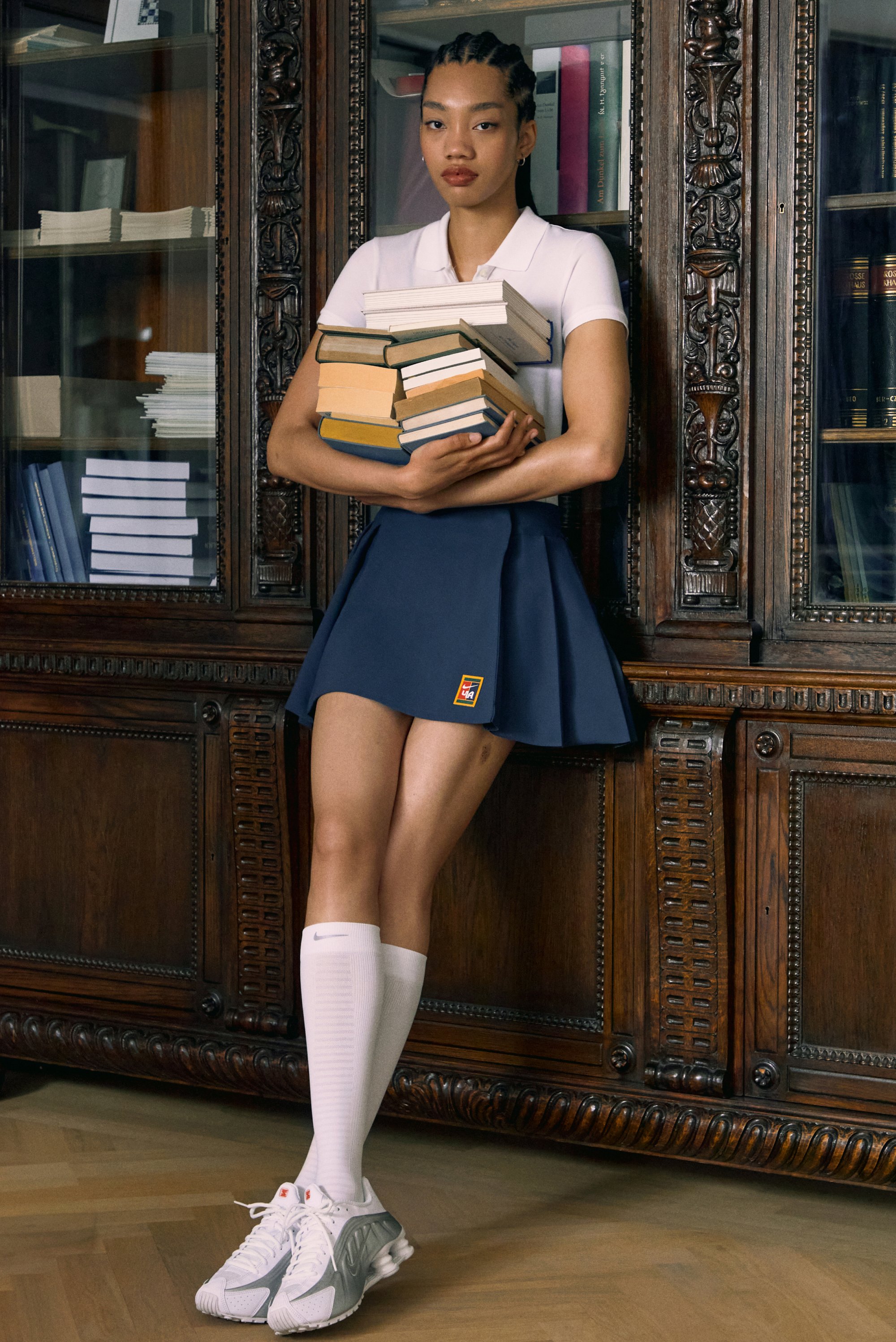 Young woman in a white polo and navy skirt holds a stack of books against an ornate wooden bookshelf in a cozy indoor setting.