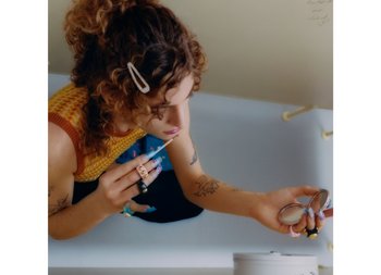 Una persona con cabello rizado se aplica maquillaje, sosteniendo un lápiz labial en una mano y un espejo compacto en la otra, rodeada de una decoración colorida.