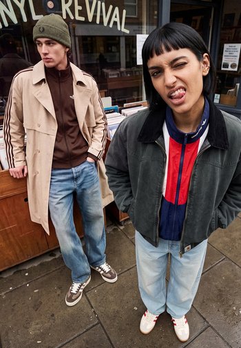 Deux jeunes adultes portant des vestes décontractées et des jeans se tiennent devant un magasin de disques vinyle, l'un faisant une grimace ludique à la caméra.