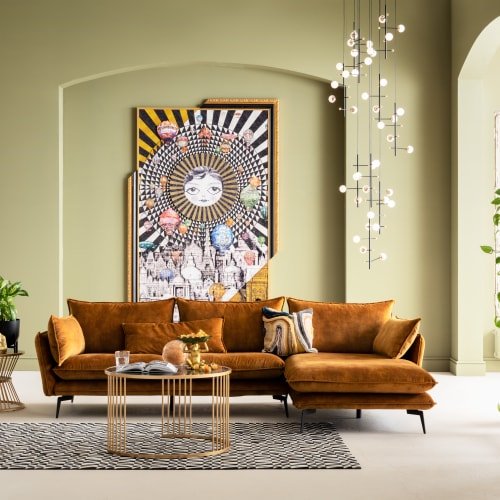 Pomarańczowa sofa w kształcie litery L z weluru, z czarnymi metalowymi nogami, ozdobnymi poduszkami oraz okrągłym stolikiem kawowym z szklanym blatem na wzorzystym dywanie. Tło w kolorze zielonym.