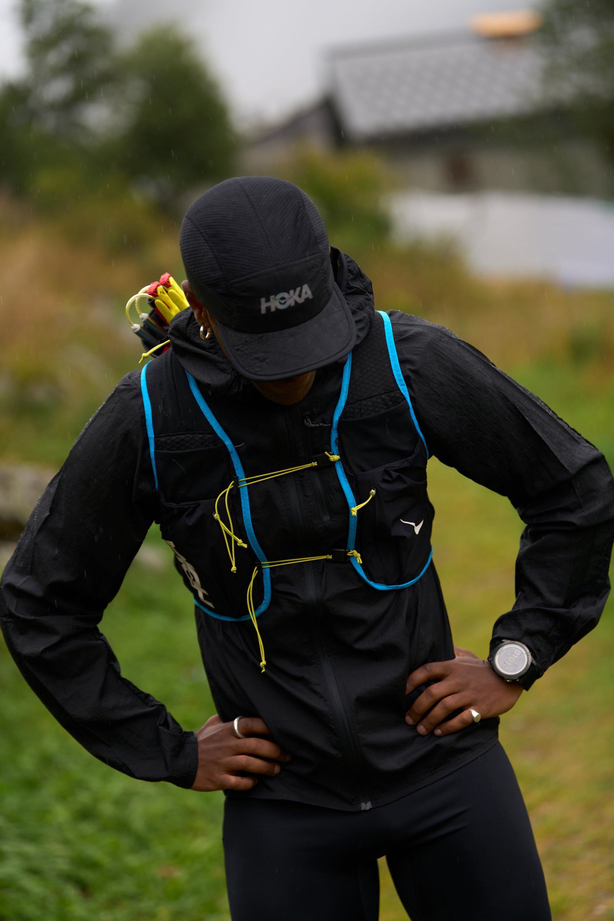 Black running vest with blue accents and yellow drawstrings, featuring mesh pockets, worn over a black shirt. Black cap with "HOKA" logo.