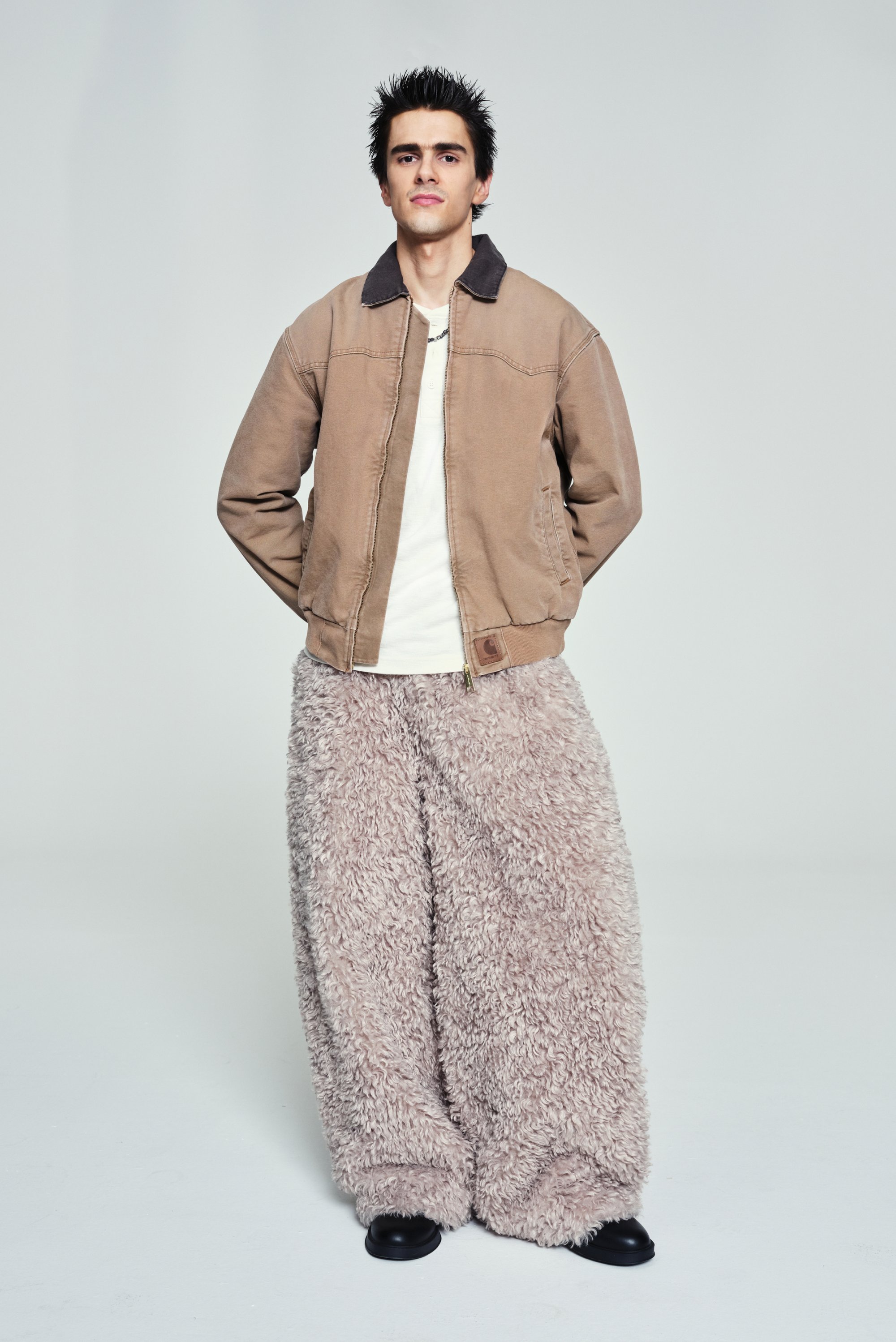 A young man stands confidently in a fluffy, light pink pant and a brown bomber jacket, against a plain background.