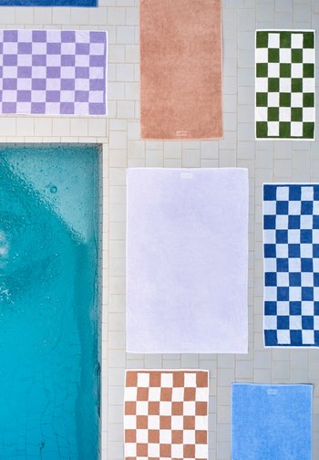 Μπλε πισίνα με πλακάκια στην άκρη, περιτριγυρισμένη από πετσέτες σε διάφορα χρώματα και καρό, οργανωμένες πάνω σε ανοιχτόχρωμα πλακάκια.