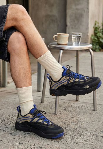 Jambes croisées portant des chaussettes blanches côtelées et des baskets The North Face bleu-noir, avec un tabouret en métal tenant une tasse en céramique et un verre d'eau à proximité.