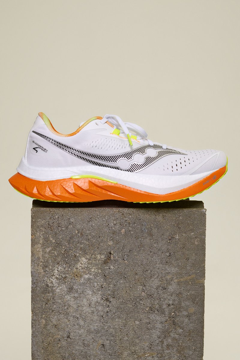 White running shoe with black accents, textured mesh upper, vibrant orange midsole, yellow laces, and reflective detailing.