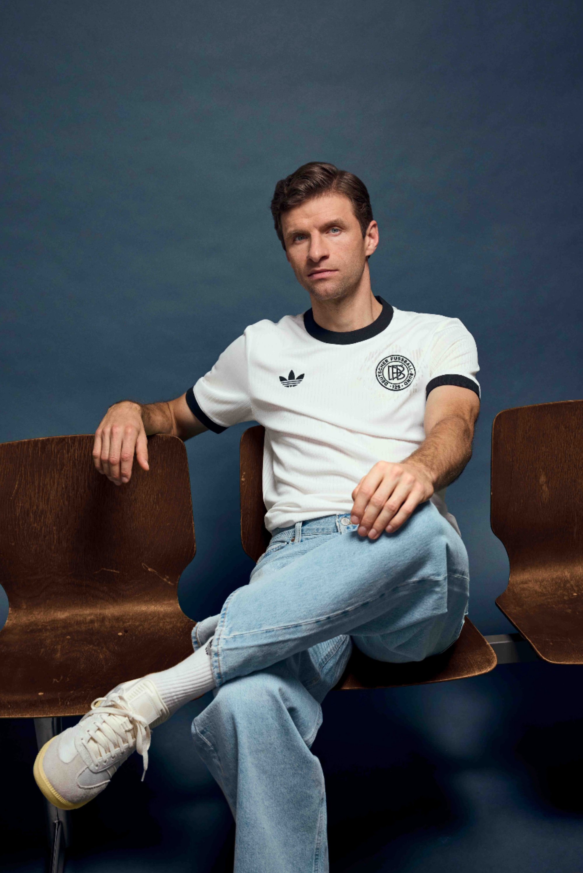 Una persona è seduta in modo informale su una sedia di legno, indossando una maglietta bianca Adidas con bordini neri, jeans azzurri chiari e sneakers bianche, su uno sfondo scuro.