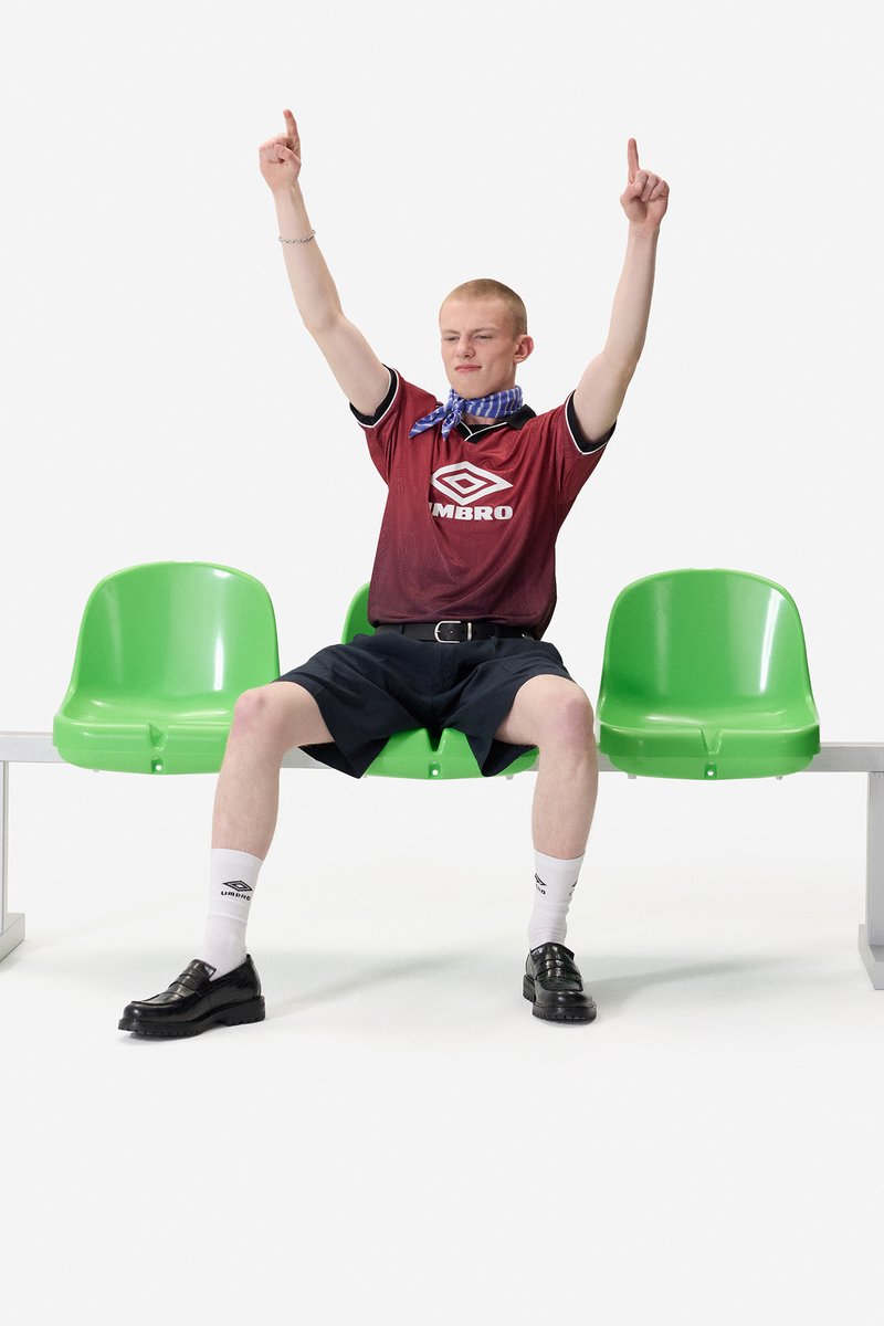 Jeune homme portant un t-shirt de sport rouge et un short noir assis sur un banc vert, levant les deux bras avec les index pointés vers le haut.