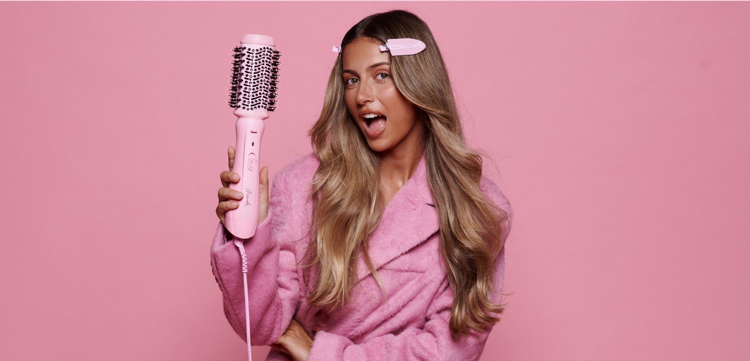 Jeune femme aux longs cheveux et pince rose tient une brosse à cheveux électrique ronde rose, portant un peignoir rose sur un fond rose.