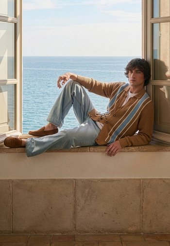 Homme en cardigan rayé marron, chemise blanche, jean bleu clair et mocassins marron assis sur un rebord de fenêtre donnant sur la mer.