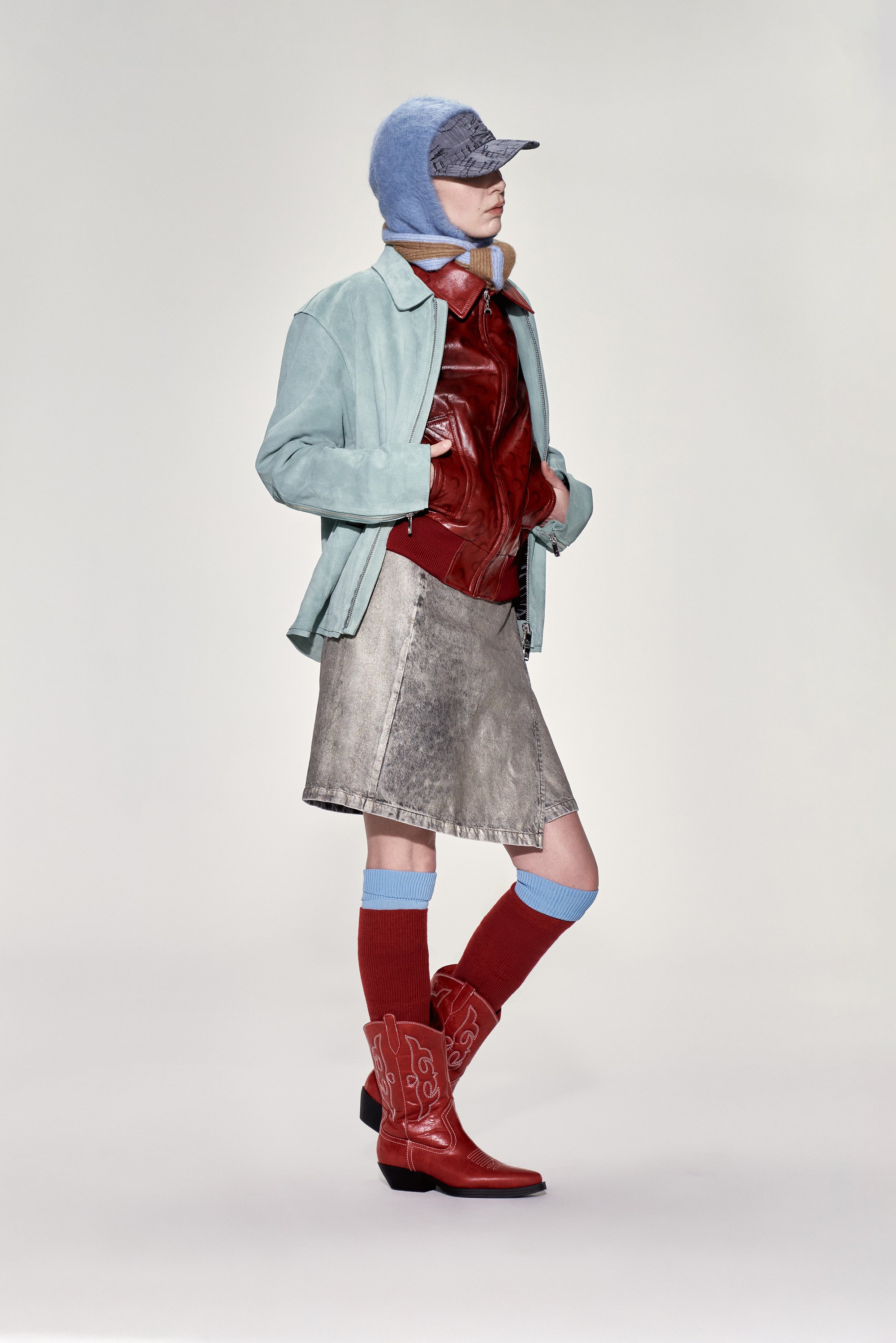 A model poses wearing a light blue jacket, red leather top, metallic skirt, and red cowboy boots with blue and red knee-length socks.