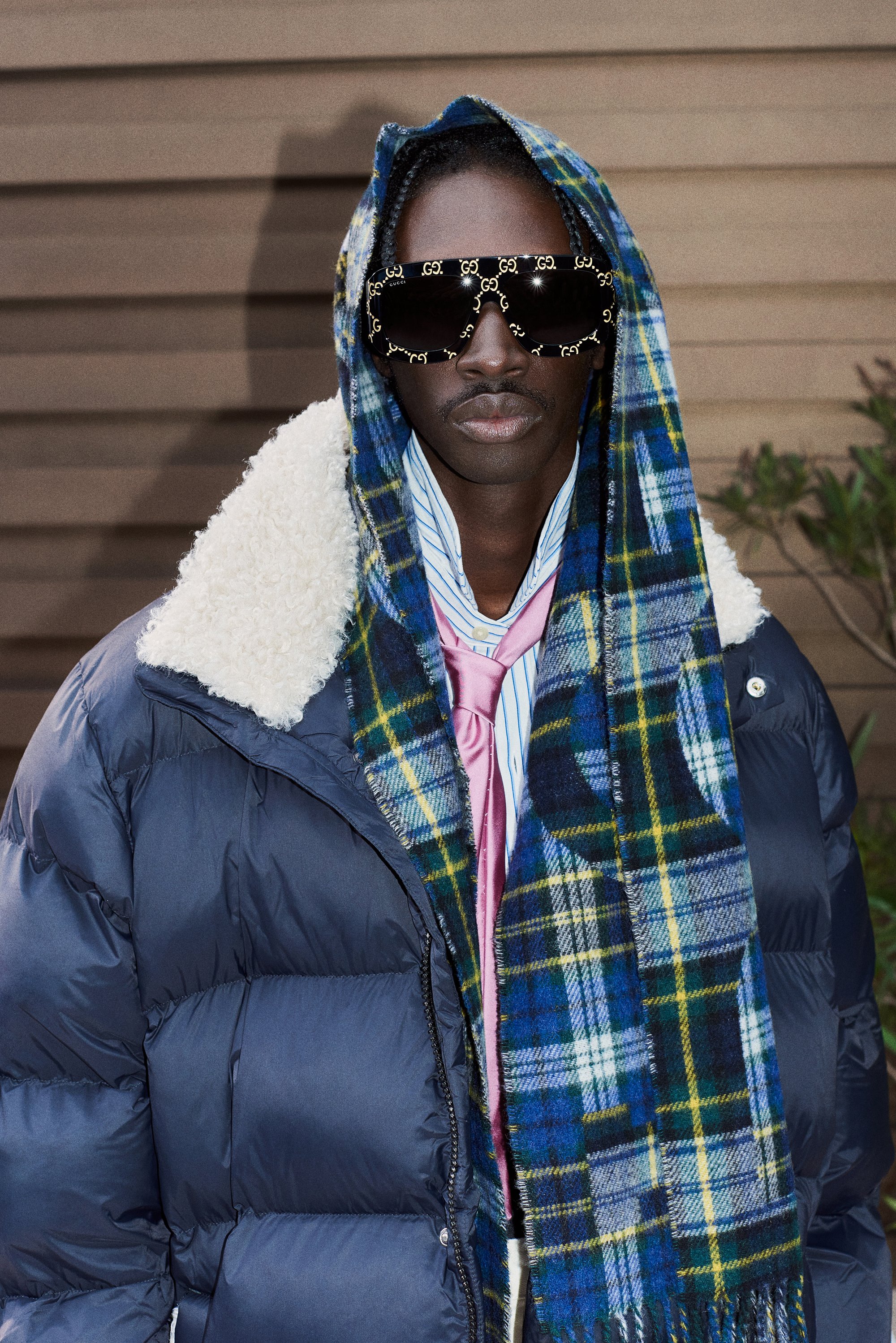 A person with dark skin wears oversized sunglasses, a puffy navy jacket, and a plaid scarf, posing against a wooden background.