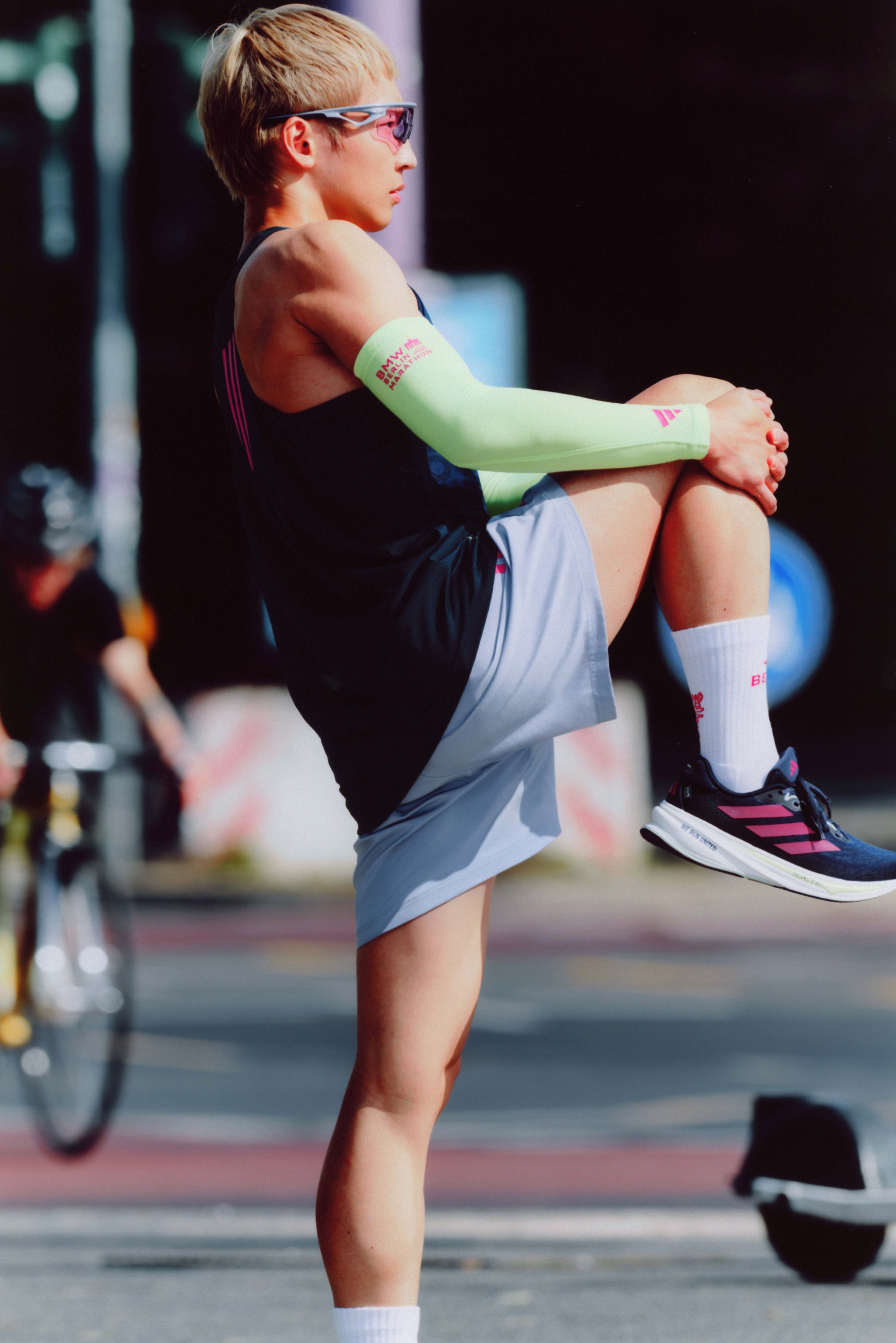 Athlete wearing a black sleeveless top, light grey shorts, green arm sleeves, and navy/pink running shoes, stretching their legs in the sunlight.
