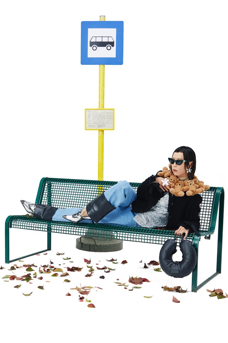 A woman with sunglasses leans against a bus stop, playfully balancing a teddy bear and a rubber tire, surrounded by colorful autumn leaves.