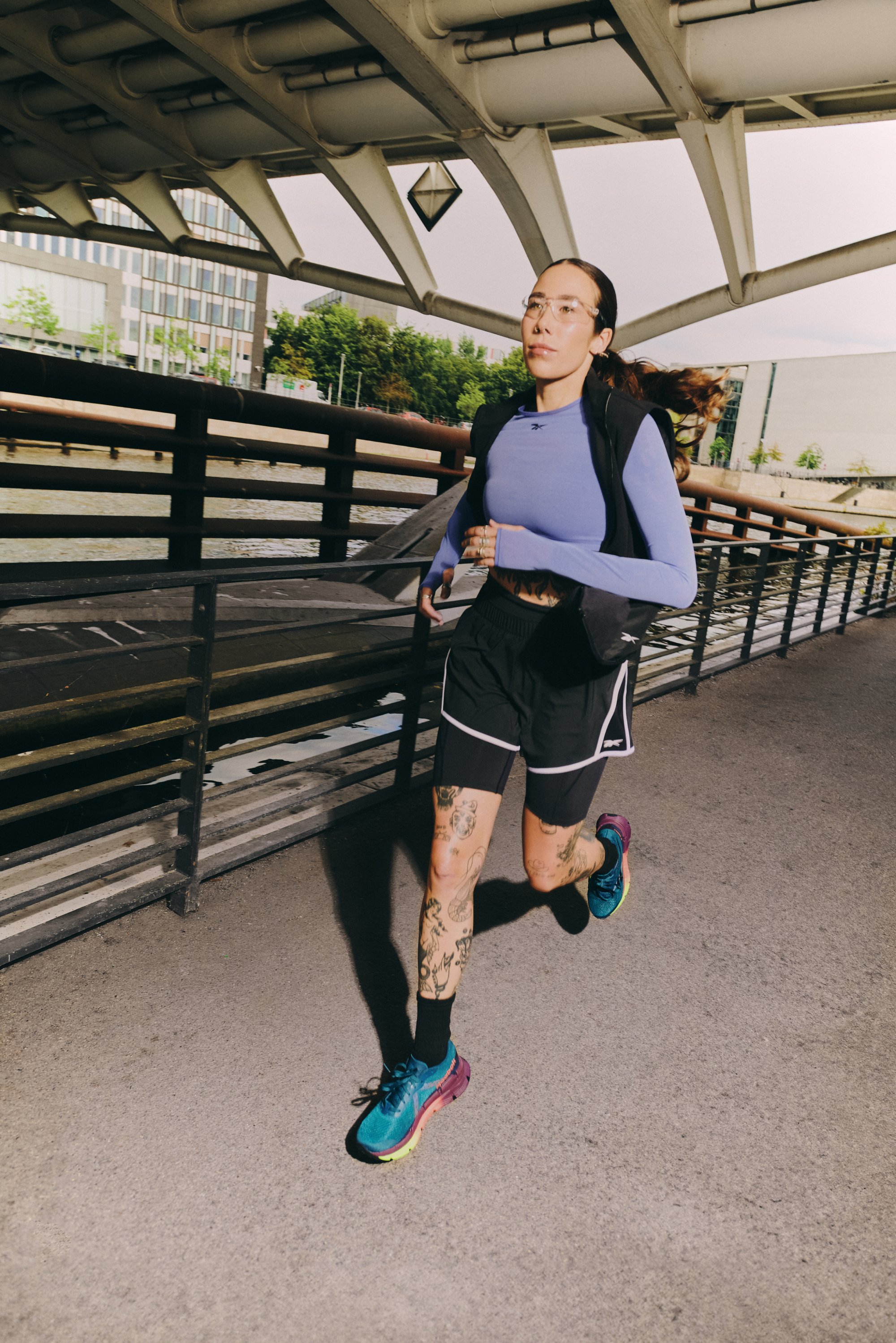 A person with tattoos runs along a riverside path, wearing a blue long-sleeve shirt, shorts, and colorful running shoes, under a bridge.