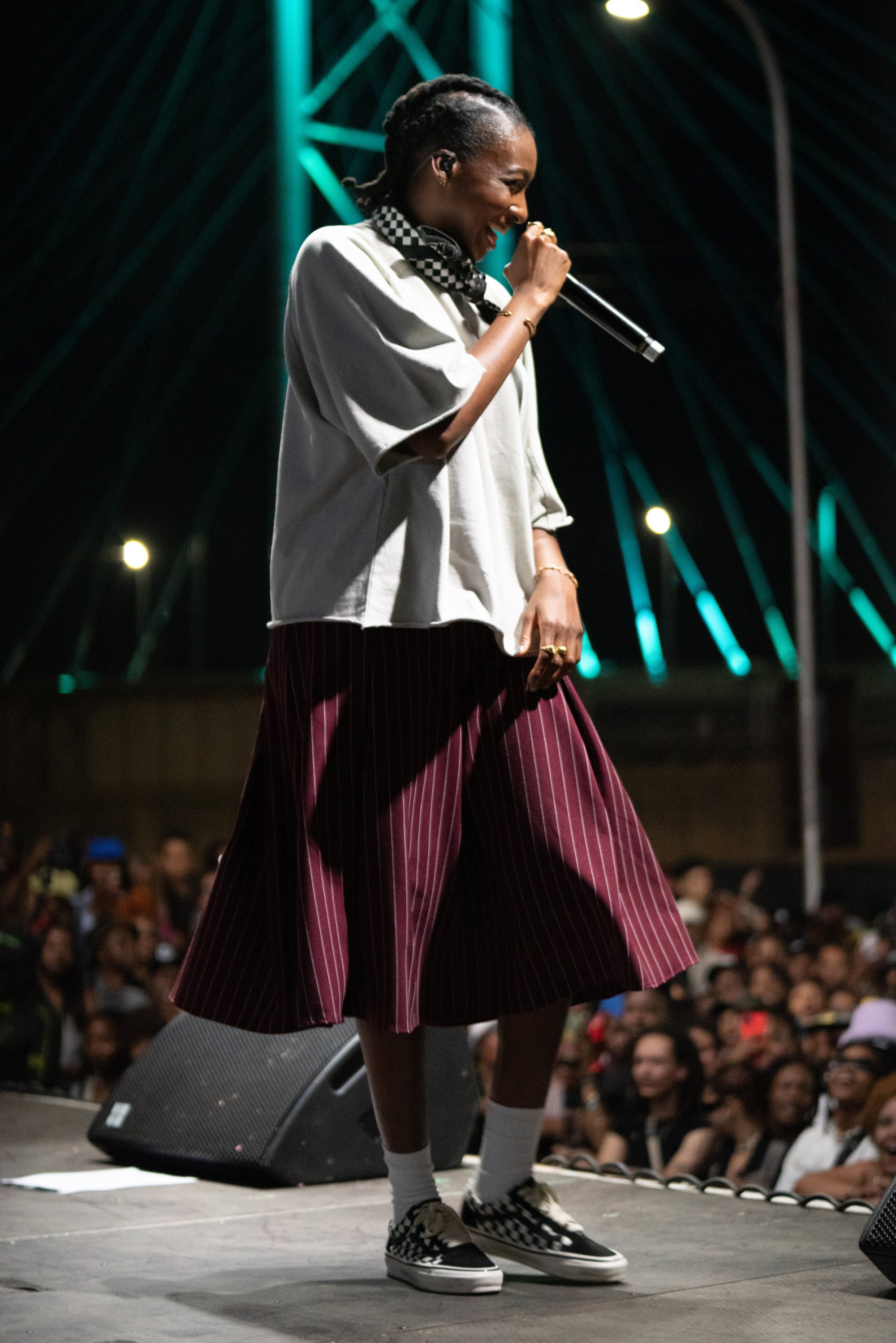 Un performer in una camicia larga e una gonna a righe canta in un microfono sul palco, con una folla vivace visibile sullo sfondo.