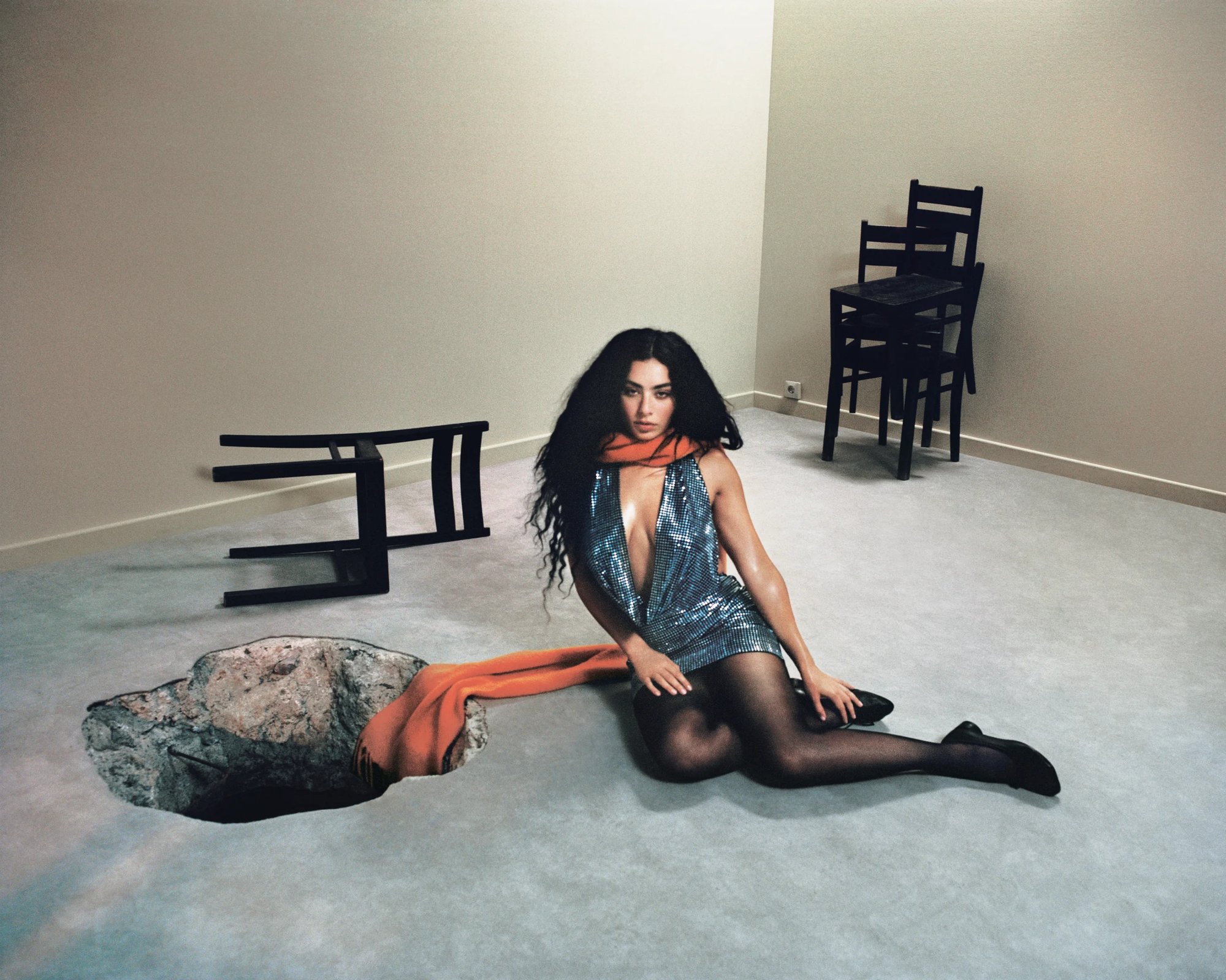 A woman in a shimmering dress and orange scarf sits near a large hole in the floor, with an overturned chair nearby in a minimalistic room.