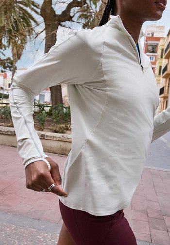 Δρομέας με λευκή μπλούζα μακριάς μανίκας με οπές για τους αντίχειρες και μαρον σορτς τρέχει σε εξωτερικό χώρο κοντά σε δέντρα και κτίρια.