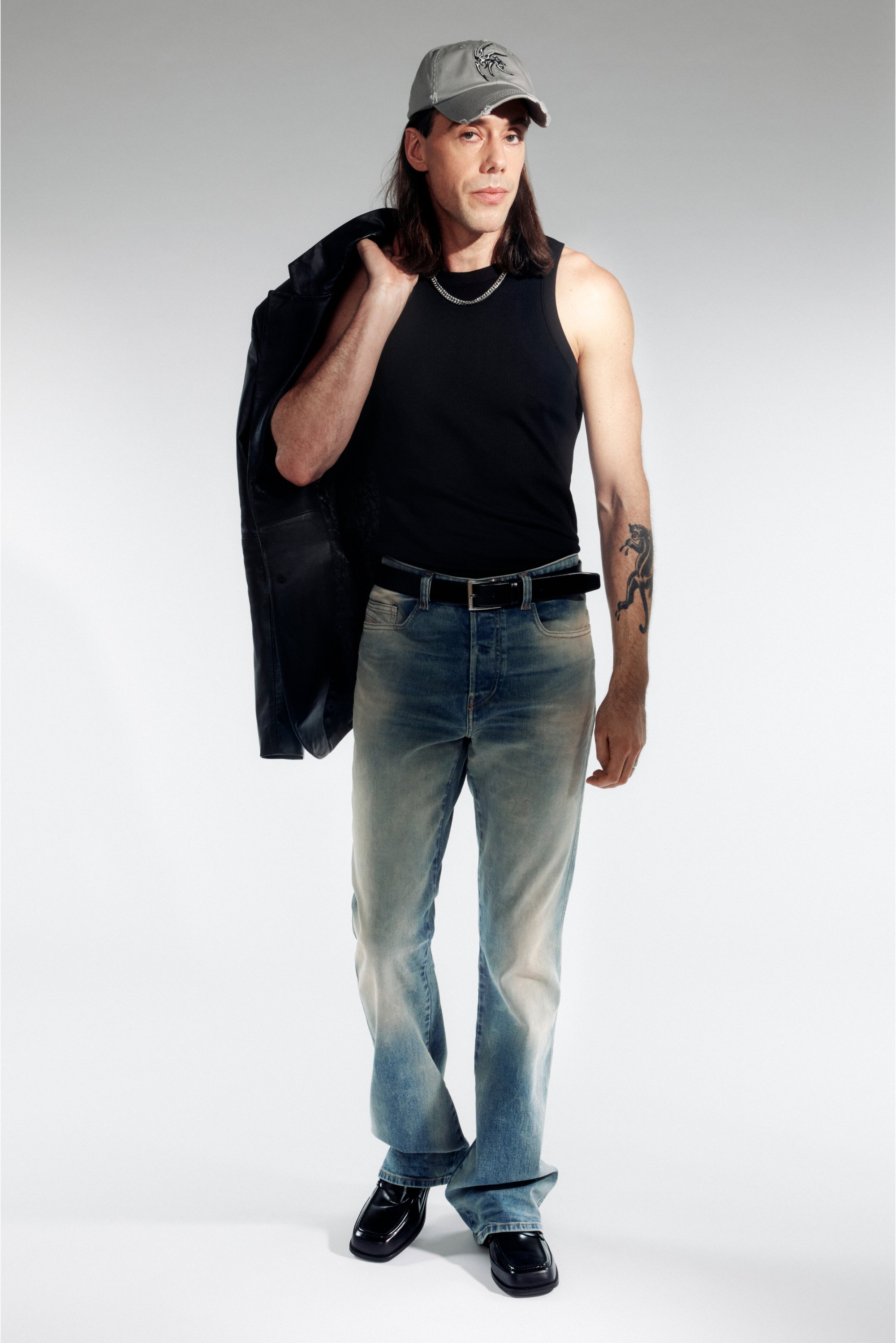 Hombre con cabello largo que lleva una camiseta sin mangas negra y jeans desgastados, sostiene una chaqueta sobre su hombro, con un fondo pálido.