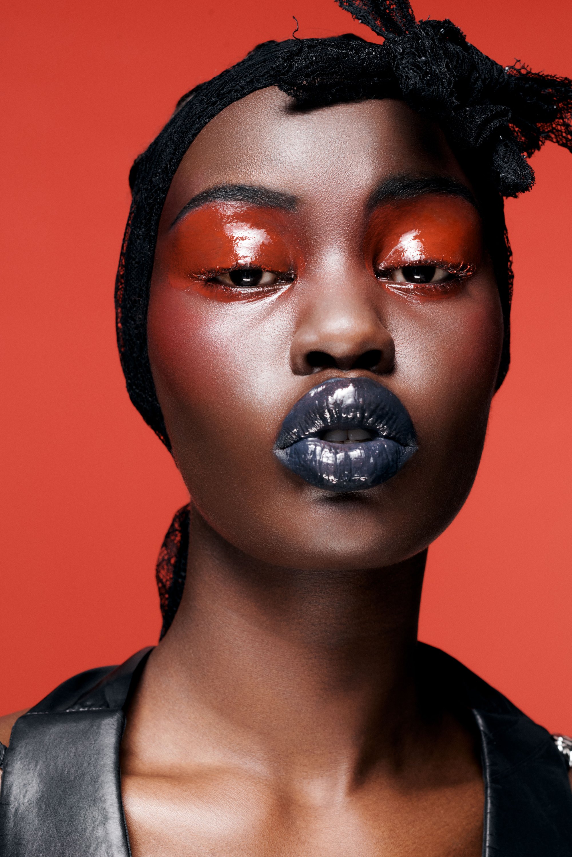 Model with dark skin and bold makeup, featuring glossy orange eyeshadow and deep blue lips, against a vibrant red background.