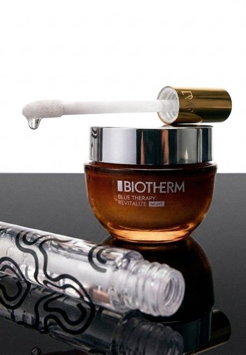 Pot de crème de nuit Biotherm Blue Therapy Revitalize avec un couvercle argenté réfléchissant et une baguette applicatrice tenant une goutte, à côté d'un tube transparent à motifs.