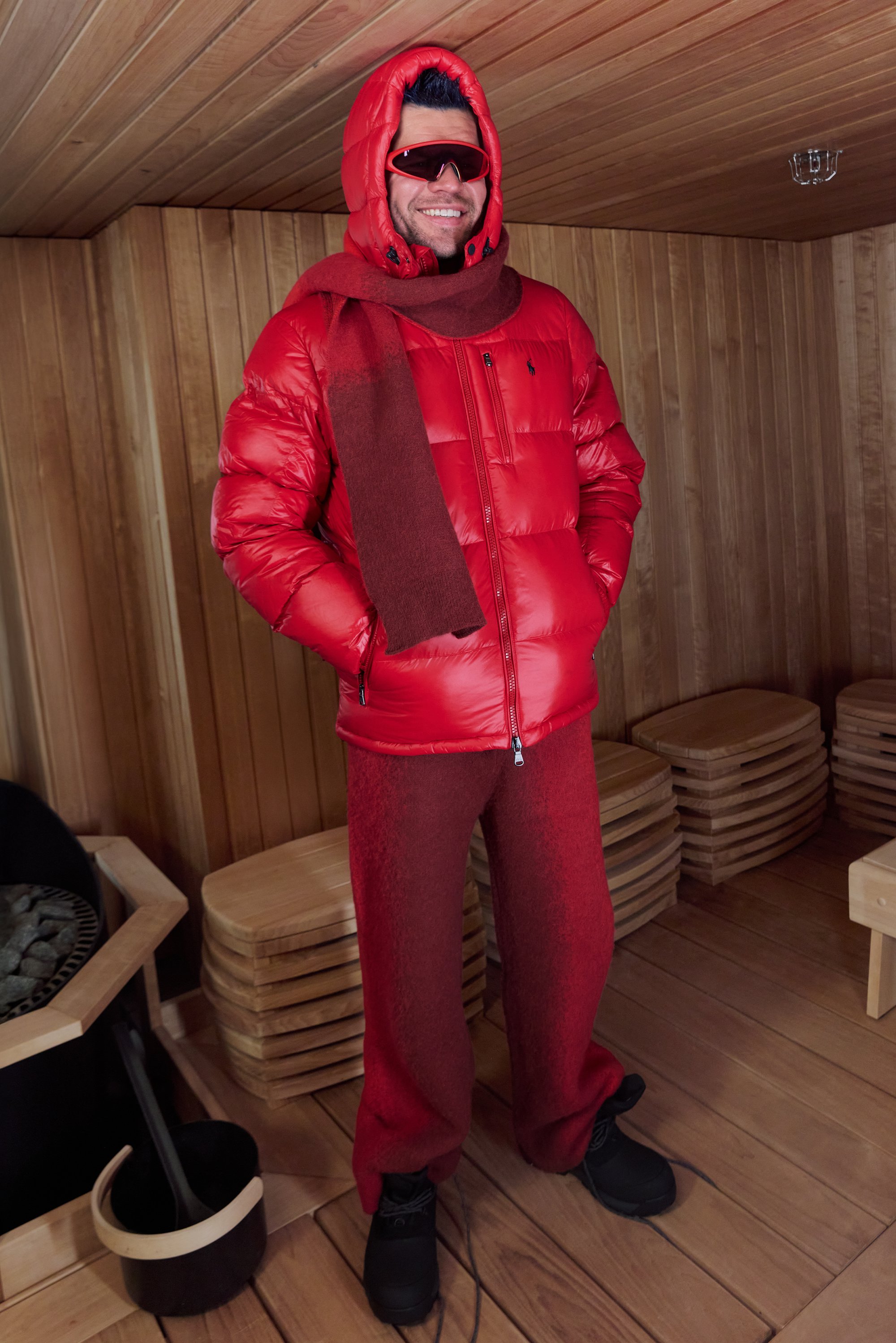 Punainen untuvatakki hupulla, yhdistetty samansävyisiin punaisiin fleecehousuihin ja villahuiviin. Mustat saappaat ja aurinkolasit täydentävät asun.