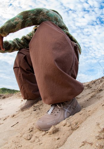 Person crouching on sandy terrain, wearing a fuzzy green jacket and brown oversized pants, with focus on light brown athletic shoes.
