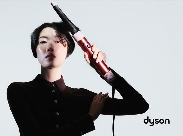 Eine Person mit kurzen Haaren hält ein rotes Dyson-Stylinggerät in der Hand, bereit, ihre Haare zu locken, und trägt ein schwarzes Shirt vor einem hellen Hintergrund.