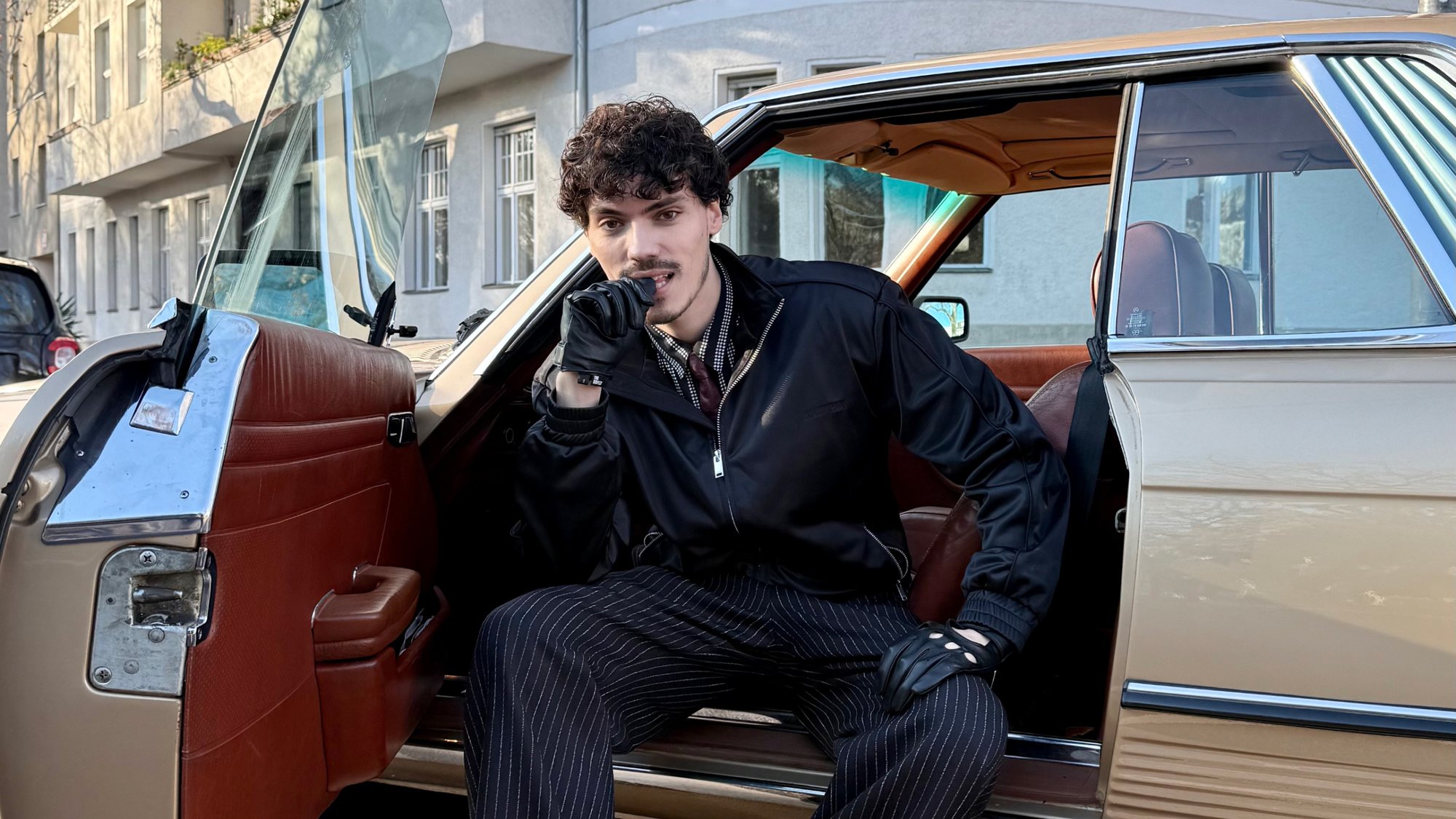Auto d'epoca con esterno crema e dettagli in cromo. Gli interni presentano sedili in pelle marrone. Una persona seduta al volante indossa guanti neri.