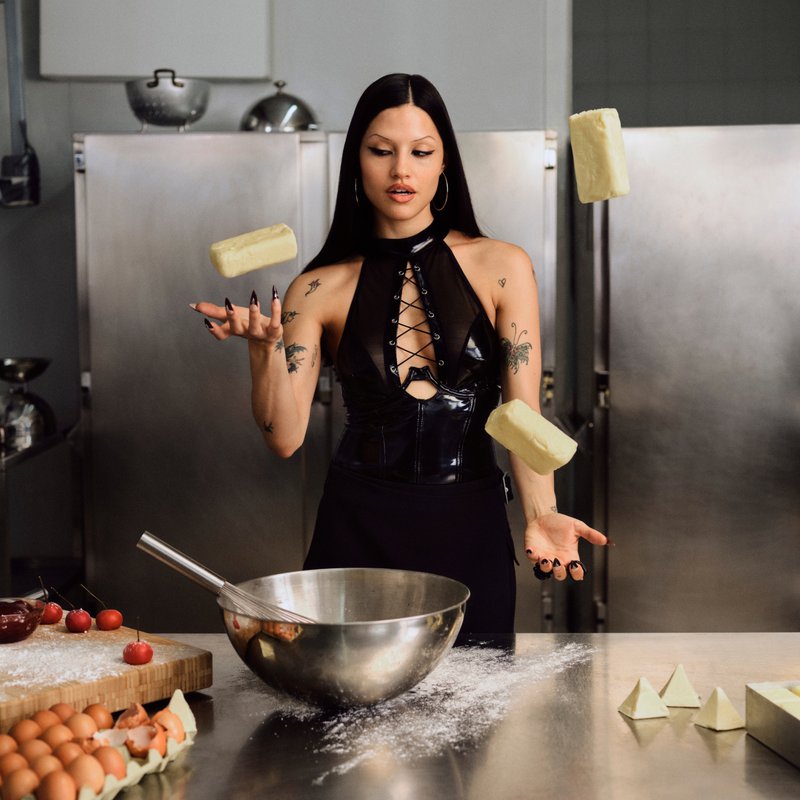 Una mujer con una blusa de cuero negra hace malabares con palos de mantequilla en una cocina, rodeada de huevos, tomates cherry y un bol de mezcla sobre una mesa de acero inoxidable.