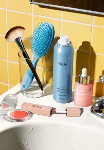 Une bombe de peinture bleue, un sérum rose, un blush transparent, et des outils incluant une brosse à cheveux bleue et un pinceau de maquillage marron sur une baignoire blanche contre des carreaux jaunes.