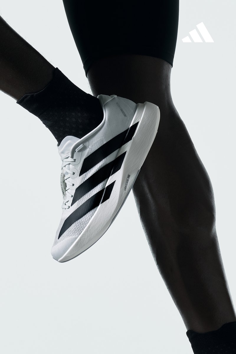 Un primo piano di una gamba che indossa una calza nera e una scarpa Adidas bianca con strisce nere, su uno sfondo minimalista e chiaro.