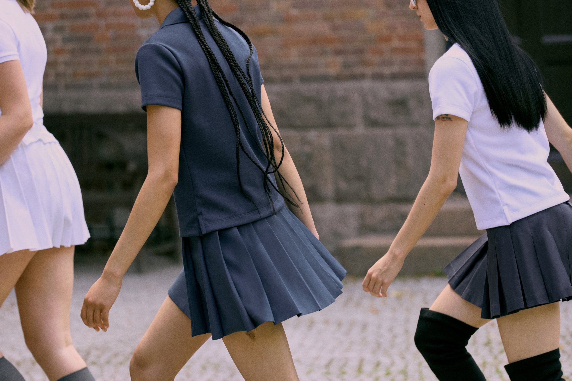 Tre individui camminano insieme, indossando gonne a pieghe corte e polo, su uno sfondo di mattoni texture.