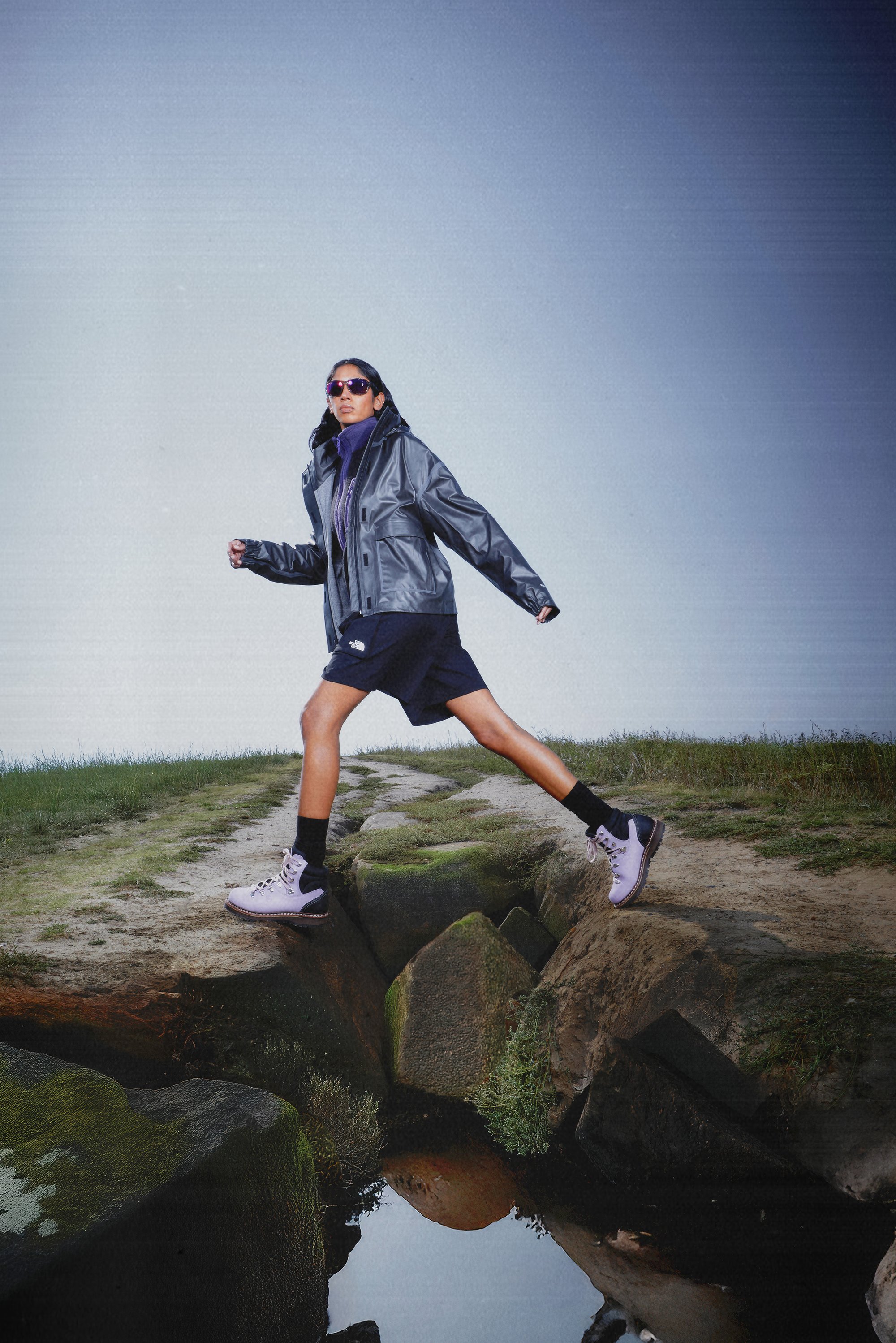 A person in a gray jacket and sunglasses strides confidently over rocks, surrounded by grass and water reflections.