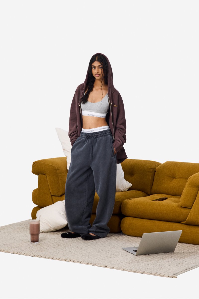 Bruine zip-up hoodie, grijze crop top en donkergrijze wijde sweatpants. Staande naast een groene bank op een beige tapijt, met een laptop en een kopje in de buurt.