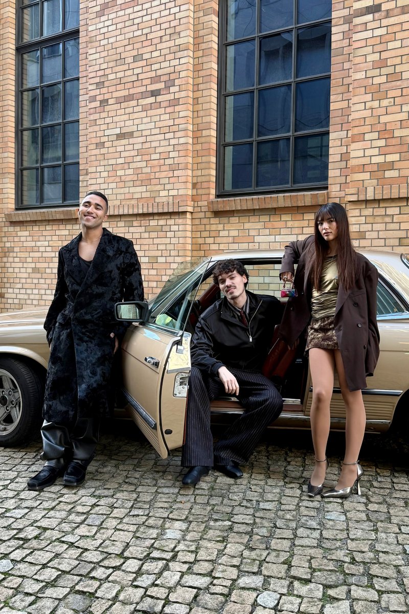 Tre persone alla moda posano di fronte a un'auto d'epoca, davanti a un edificio in mattoni. Una sorride, mentre l'altra si appoggia casualmente alla porta.