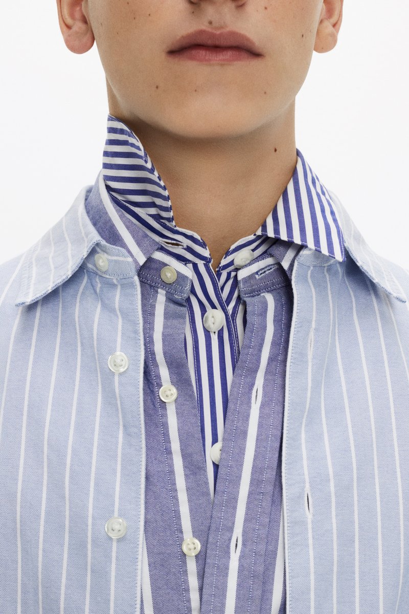 A young person wears a layered shirt with blue and white stripes, featuring a large collar and buttons visible down the front.