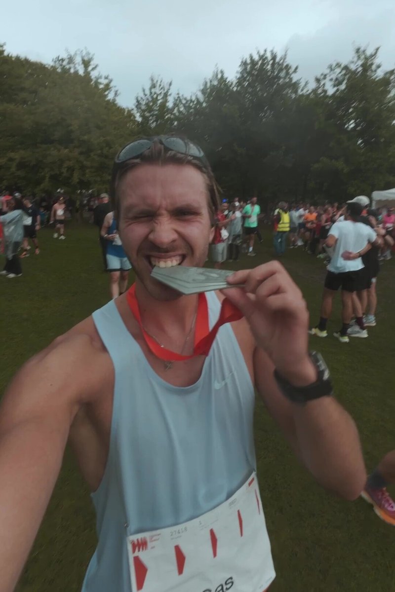 Athlet in einem hellblauen Tanktop beißt in eine Silbermedaille mit rotem Band. Eine Startnummer ist auf einer Brustnummer am Bauch zu sehen. Grüner Park im Hintergrund.