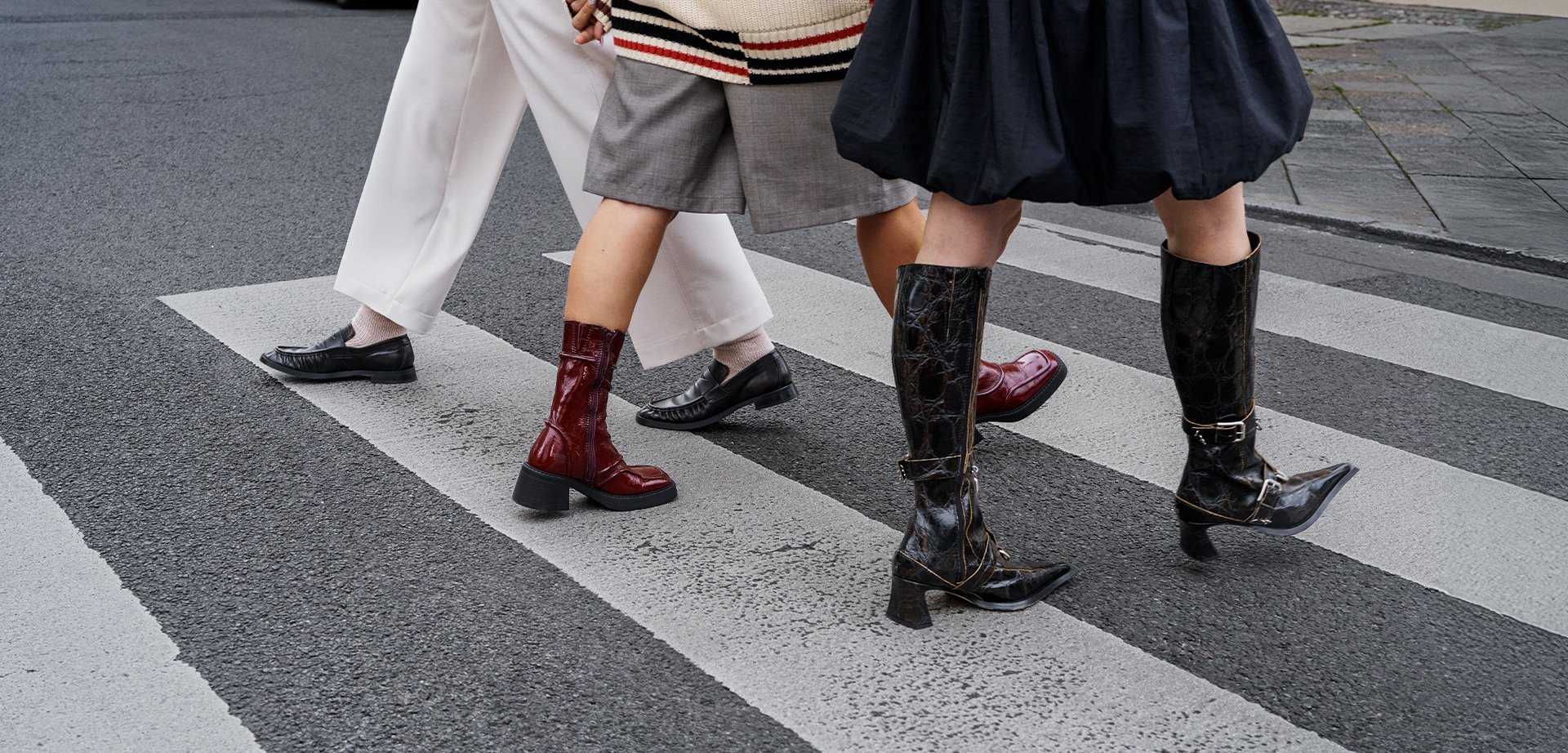 Three pairs of boots crossing a street: shiny red ankle boot, black pointed knee-high with buckle, and black textured low-heeled shoe.
