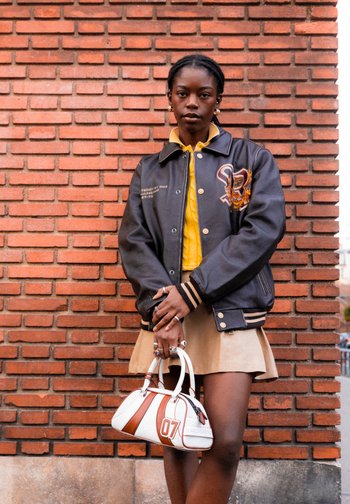 Eine Person steht vor einer Ziegelwand, trägt eine schwarze Jacke über einem gelben Shirt und einem beigen Rock und hält eine weiße Handtasche mit der Aufschrift '07'.