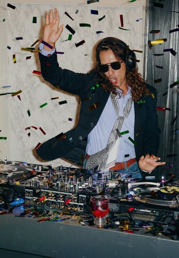 DJ ved en platespiller med svarte hodetelefoner, iført svart jakke, blå stripete skjorte og mønstret slips. Fargerikt konfetti sprer seg rundt.