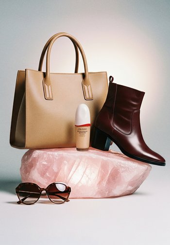 Brązowa torebka ze skóry, brązowy skórzany botek na różowym kamieniu, podkład w butelce oraz okulary przeciwsłoneczne w oprawkach w panterkę na białej powierzchni.