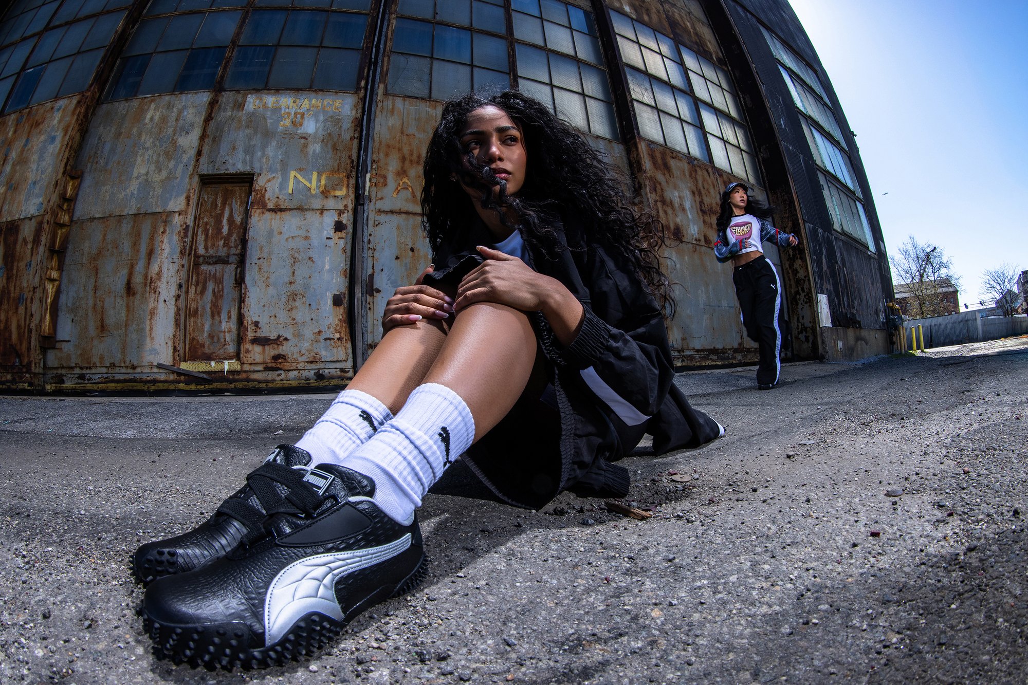 Una joven con cabello rizado se sienta en el suelo, mostrando zapatillas Puma, mientras otra mujer camina con ropa deportiva casual en un ambiente industrial.