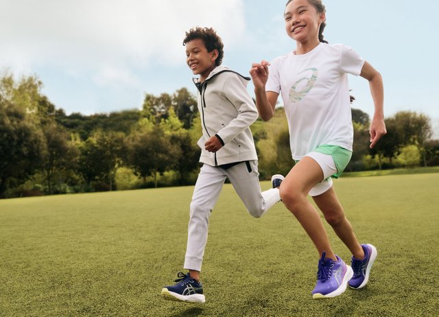 To barn som løper på gressplen utendørs, smilende og iført sportsklær og joggesko, med trær og en blå himmel i bakgrunnen.