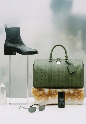 Czarny skórzany botek za kostkę na przezroczystej podstawce, zielona torba typu duffel z wzorem na kryształowej szczotce, czarny dezodorant Armani Code oraz srebrne okulary przeciwsłoneczne na białej powierzchni.