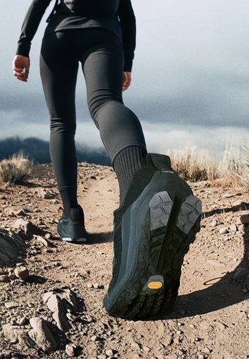 Scarpe da ginnastica nere con una tomaia in rete testurizzata, suola robusta e un motivo di battistrada distintivo, mostrate su un sentiero con terreno roccioso.